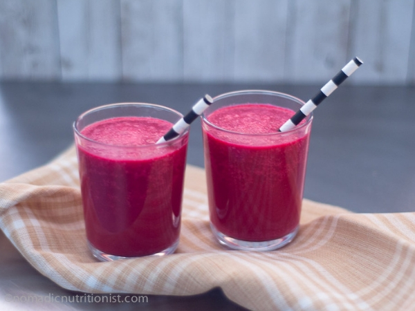 Two glasses filled with red beet shake and a black & white stripped paper straw on a tan and white tea towel.