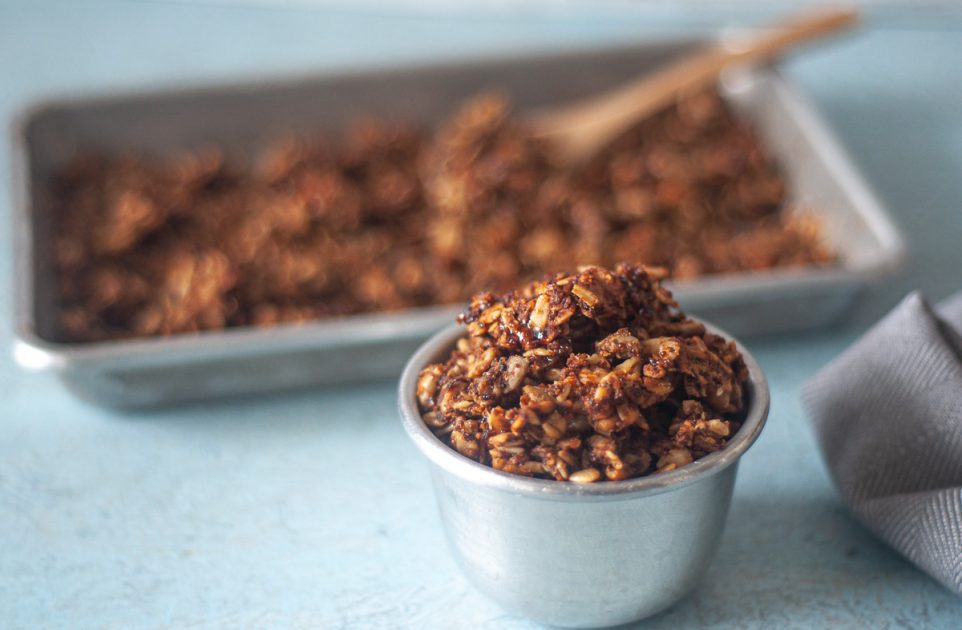 A small silver dish of peanut butter kaniwa granola with a sheet pan of granola in the background