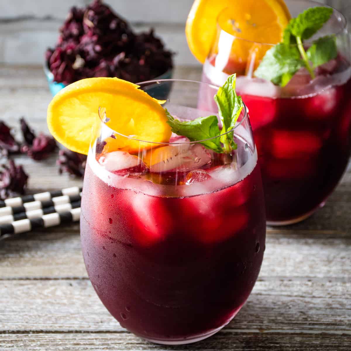 A glass of cold sorrel with ice, a sprig of mint and slice of orange sits on a wooden countertop. A second glass of the red sorrel sits behind with black & white paper straws and dried sorrel in a turquoise bowl.