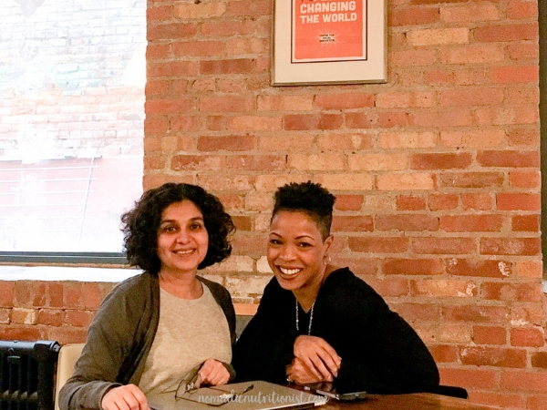 Two women, both with brown skin and short dark hair, sitting in front of a closed laptop, a pair of glasses sitting on the laptop. Both women are smiling and looking at the camera.