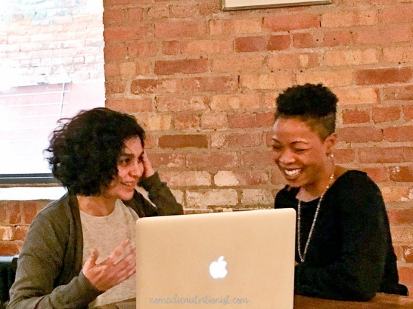 Two women, both with brown skin and short dark hair, sitting in front of a laptop. One woman is gesturing and one is laughing