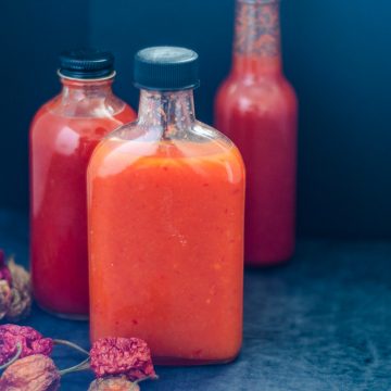 A rectangle bottle of orange-coloured homemade lactofermented pepper sauce with two bottles of red pepper sauce in the background.