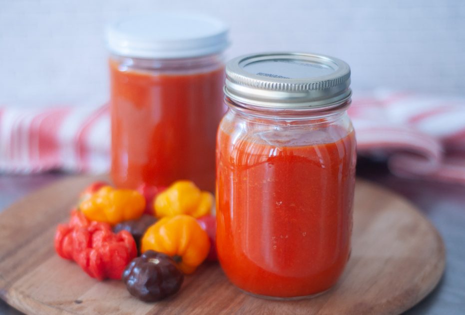 Two jars of lactofermented pepper sauce on a wooden board with fresh habanero peppers beside them.