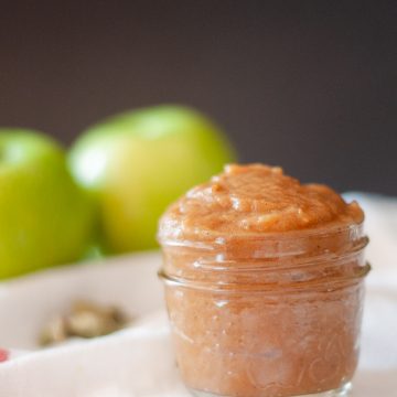 A small glass jar with apple butter. In the background are two green apples.