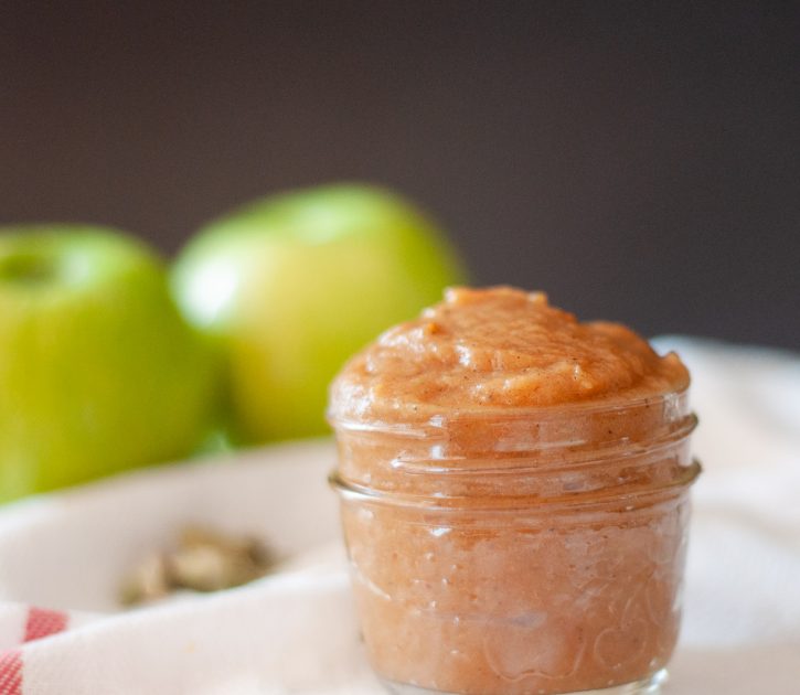 A small glass jar with apple butter. In the background are two green apples.