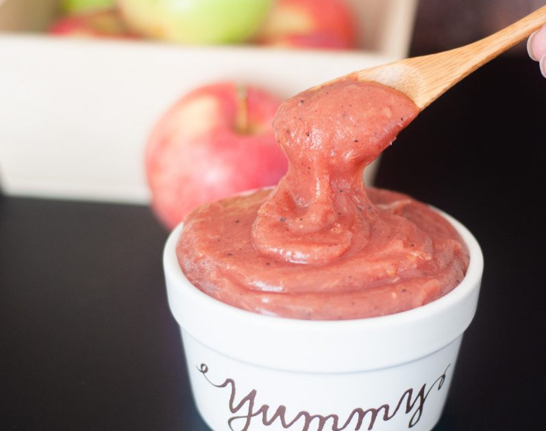A small white bowl with the word 'yummy' on the front filled with apple butter. A small wooden spoon is scooping some out.