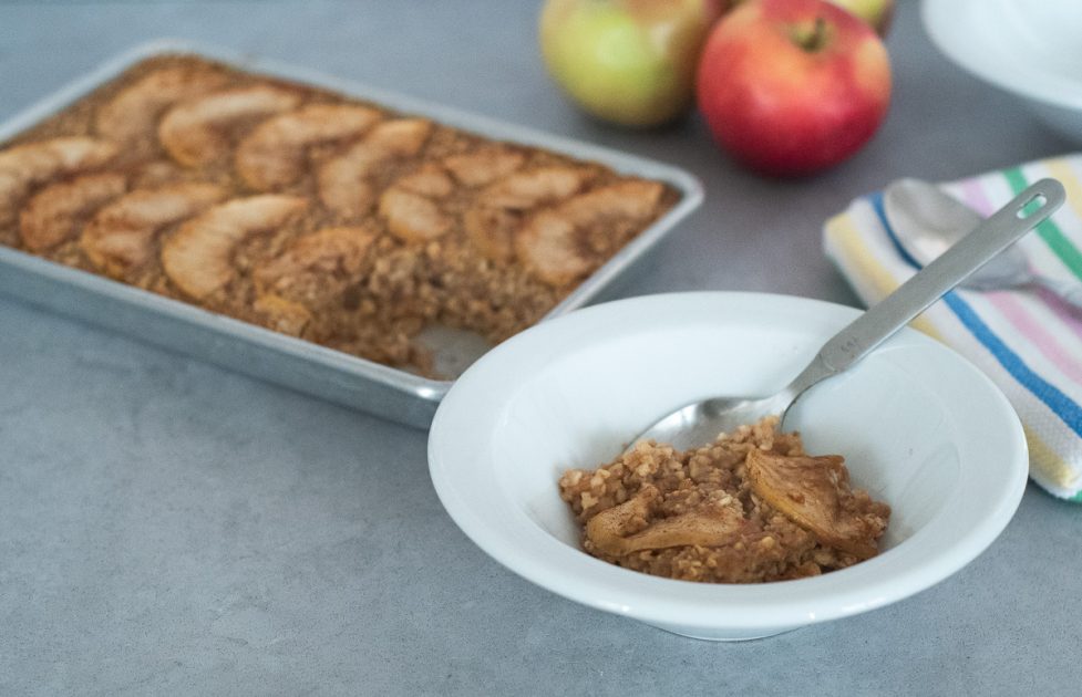 A white bowl of apple cinnamon baked oatmeal with a small sheet pan of oatmeal in the background