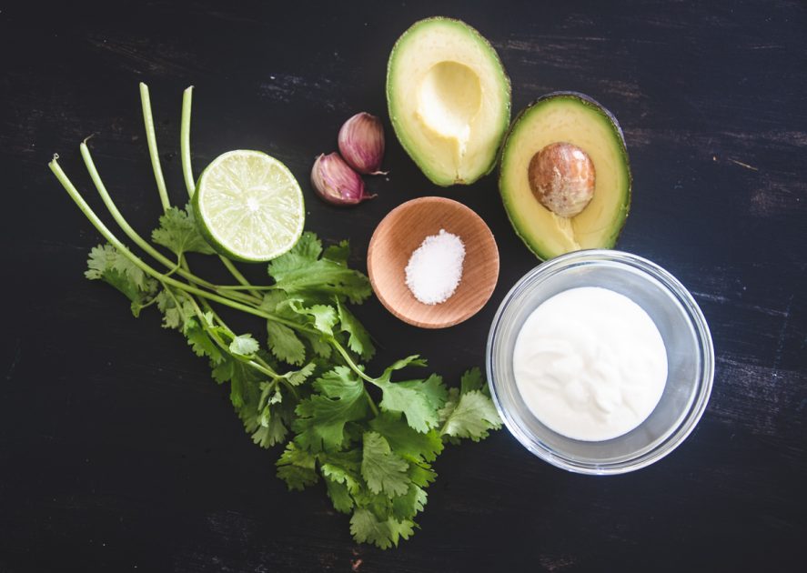 Ingredients for Avocado Cilantro Cream: A small bunch of fresh cilantro, half a lime, 2 garlic cloves, salt and coconut milk on a black table top.