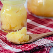 A wooden spoonful of ghee with a small jar of ghee in the background, sitting on a red & white checkered napkin.