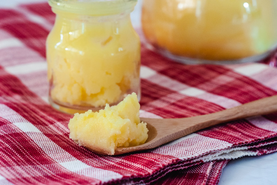 A wooden spoonful of ghee with a small jar of ghee in the background, sitting on a red & white checkered napkin.