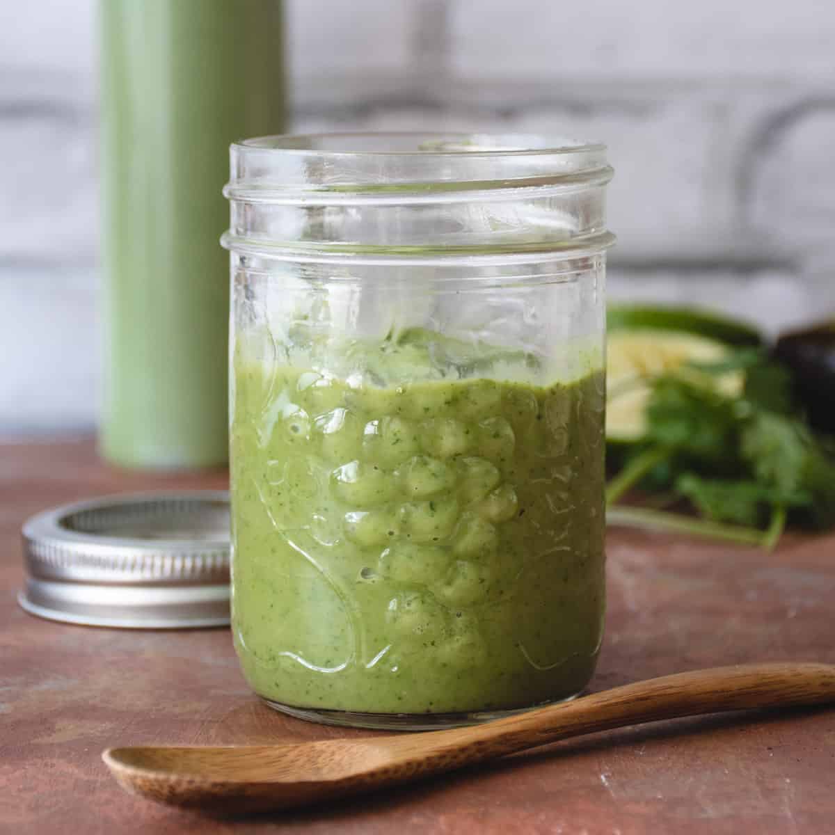 A small jar half filled with green avocado cilantro cream. A small wooden spoon sits in front of the jar.