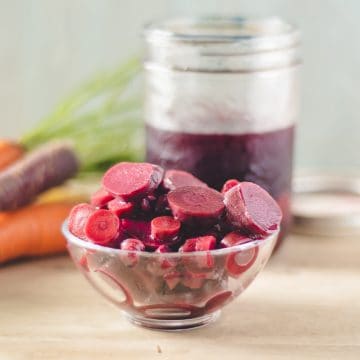A small bowl of lactofermented slices of purple carrots. Raw whole carrots in the background.