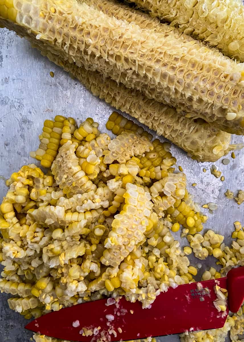 Fresh corn kernels just cut off the cob on a sheet tray. The empty corn cobs are at the top of the picture A red knife sits in the pile of corn,