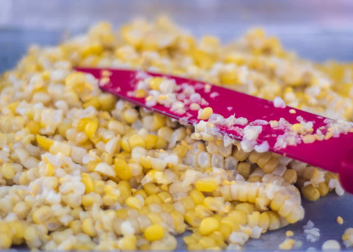 Fresh corn kernels just cut off the cob. A red knife sits in the pile of corn,