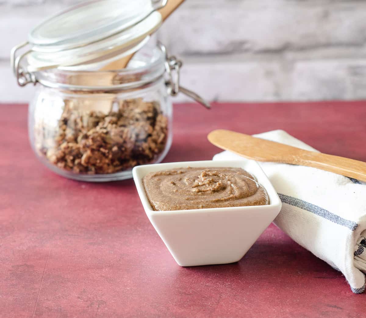 A small white dish with granola butter. A wooden knife sits on a white and blue napkin. A jar with granola is in the background