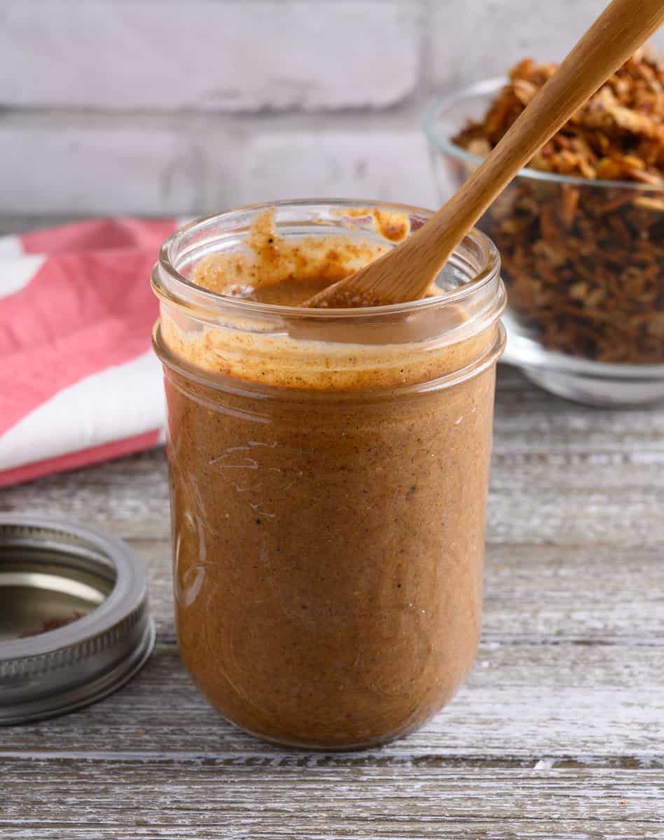 A jar of granola butter with a wooden spoon in the jar. A small bowl of granola is in the background.