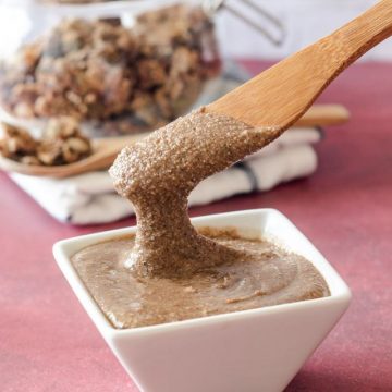 A small white dish with granola butter, above it is a wooden knife scooping out some granola butter. A jar with granola is in the background