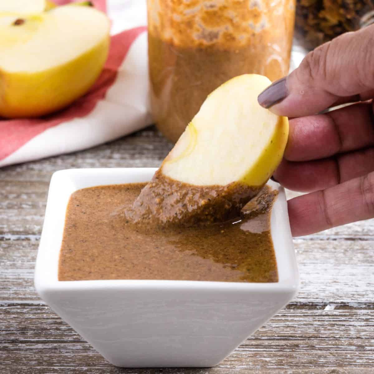 A small square bowl filled with granola butter. A hand with an apple slice is being dipped into the granola butter.