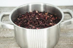 A small metal sauce pot filled with water and sorrel (hibiscus) flowers floating on top sitting on a wooden counter top.