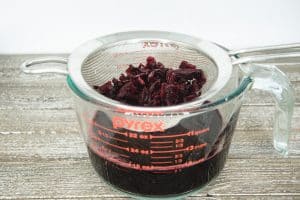A glass Pyrex measuring cup topped with a small wire strainer. In the strainer are red hibiscus flowers and inside the cup is the red hibiscus juice.