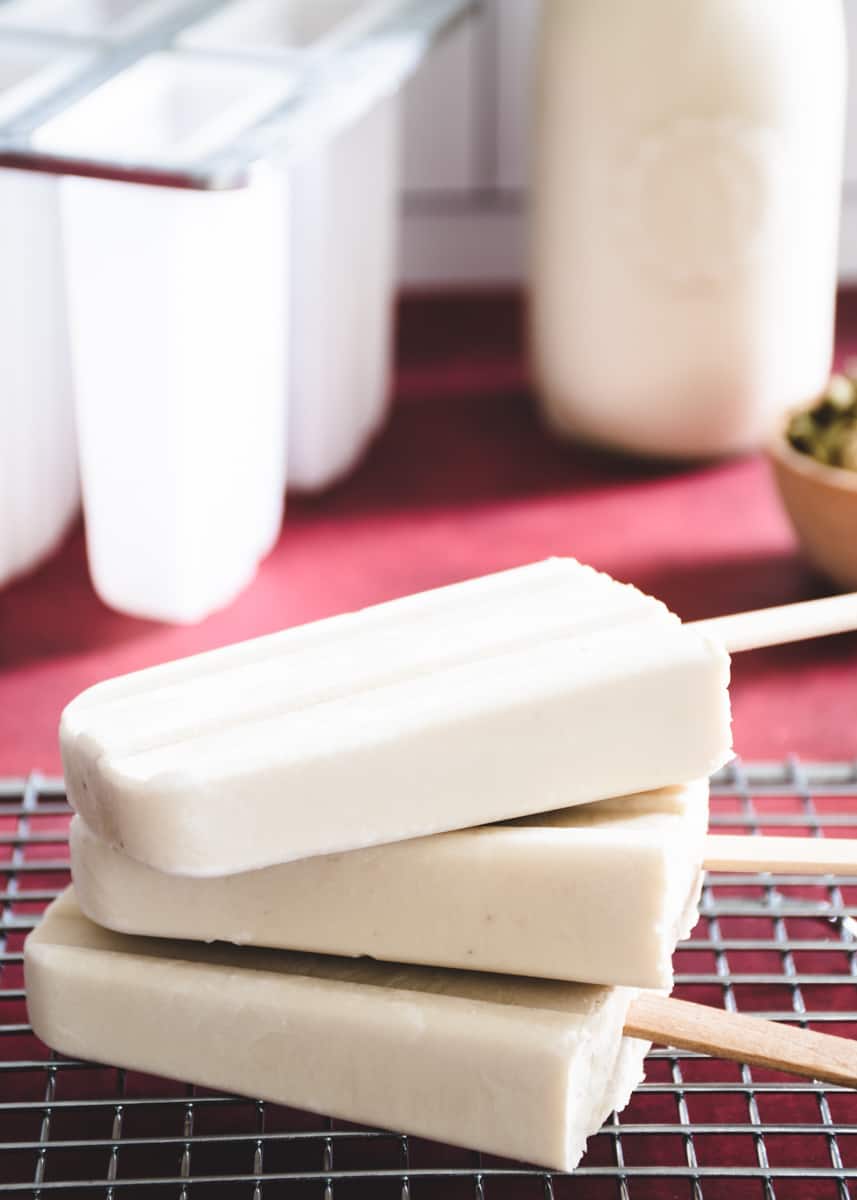 3 off-white coconut cardamom popsicles stacked on a wire rack on a red surface. In the back ground is a small dish of cardamom pods and a bottle of coconut milk.