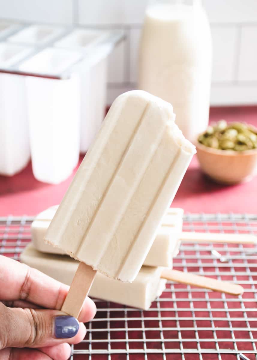 A hand holding an off-white coconut cardamom popsicle with a bite taken out of it. In the background are 2 popsicles on a wire rack, a small dish of cardamom pods, a bottle of coconut milk and a plastic popsicle mold on a red surface.