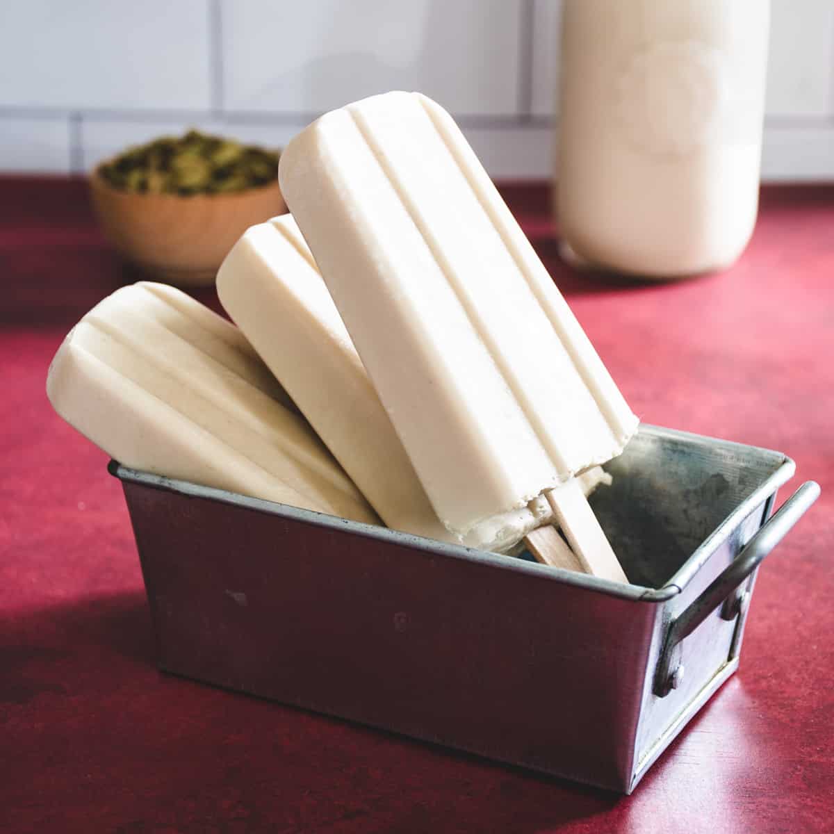 3 off-white coconut cardamom popsicles in a steel dish on a red surface. In the back ground is a small dish of cardamom pods and a bottle of coconut milk.