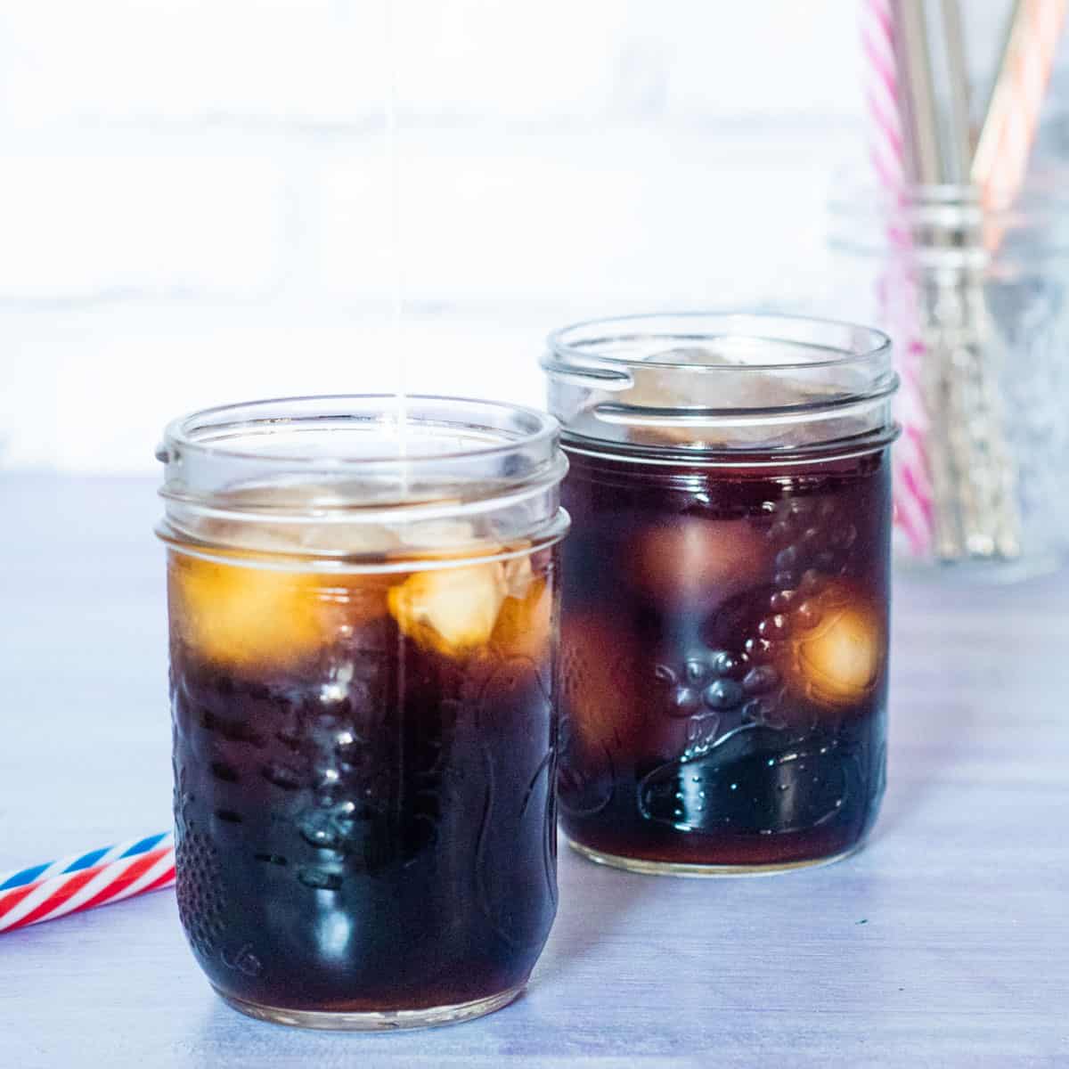 2 glass jars of black cold brew coffee with ice. Two striped straws (red & white and blue & white) are beside the jars and a cup with more colourful straws are in the background.