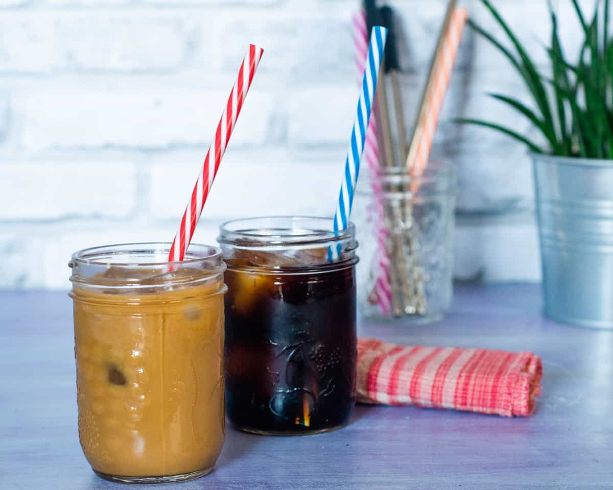2 glass jars of cold brew coffee with ice - one black and with cream. A colourful striped straw is in each jar (red & white and blue & white).In the background is a small green plant, a red & white cloth napkin and a jar with more straws.