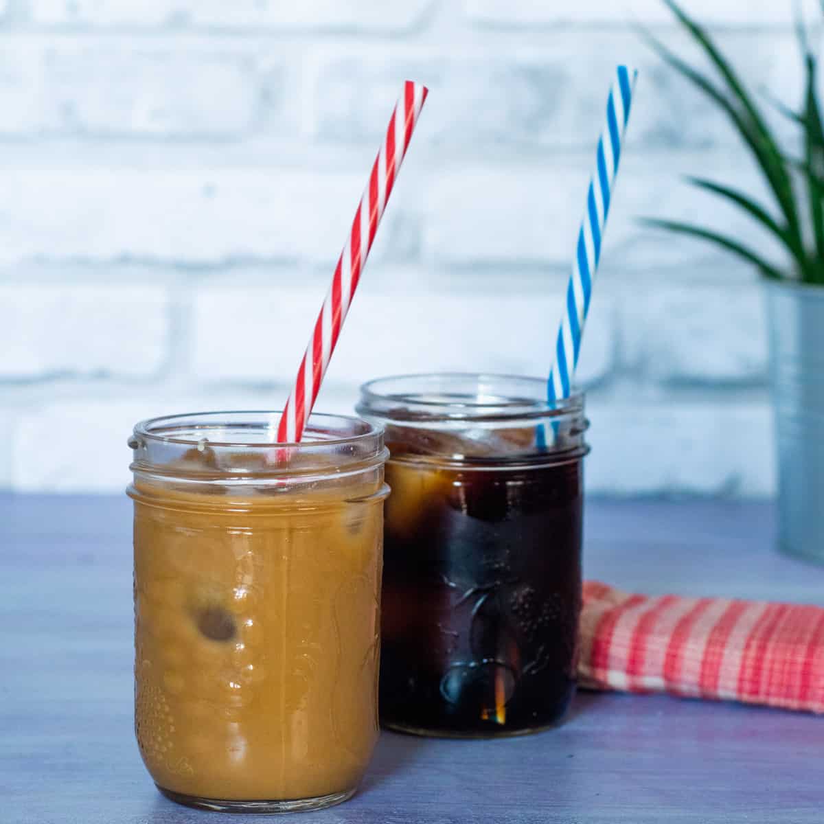 2 glass jars of cold brew coffee with ice - one black and with cream. A colourful striped straw is in each jar (red & white and blue & white).In the background is a small green plant and a red & white cloth napkin.