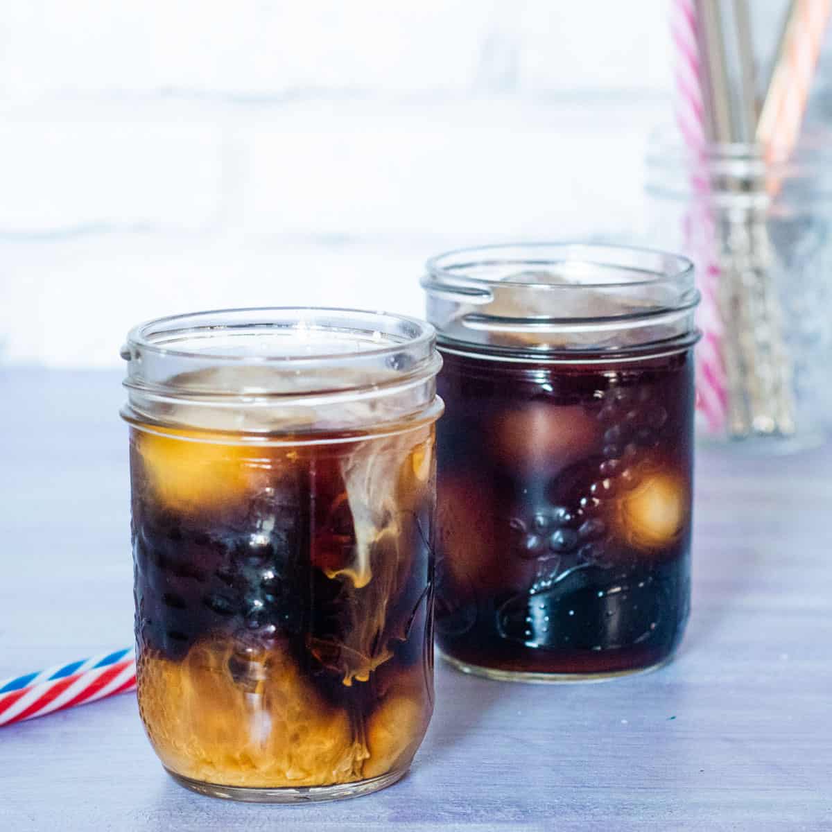 2 glass jars of cold brew coffee with ice - one black and with cream. A colourful striped straw is in each jar (red & white and blue & white).In the background is a jar with more straws.
