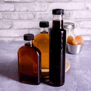 3 glass bottles of homemade simple syrup of varying colours: dark brown, amber and golden on a grey surface. In the background is a small metal dish with pinkish rock sugar and a glass jar of white sugar.