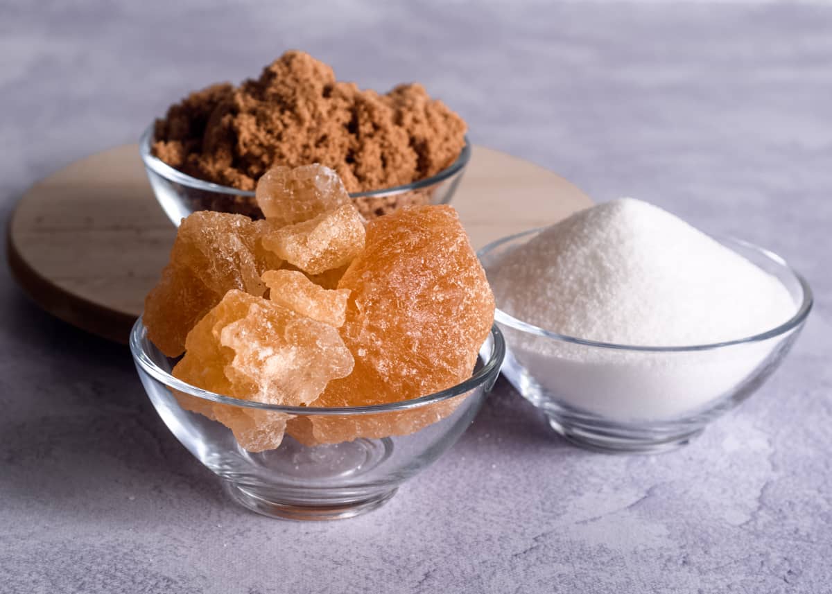 A close up of 3 small glass bowls with different kinds of sugar: pinkish rock sugar, white sugar and in the background dark brown sugar.