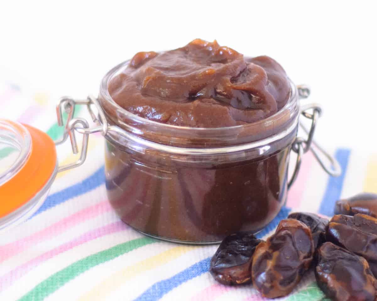 A small glass jar with the lid open filled with date paste on a colourful striped surface. A few whole dates sit beside the jar.