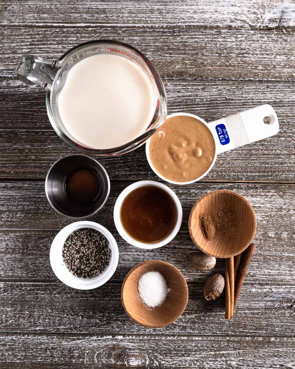 Overhead shot of peanut punch ingredients on a wooden table top. Milk, peanut butter, vanilla, honey, cinnamon, salt, chia seeds, whole nutmeg and cinnamon sticks.