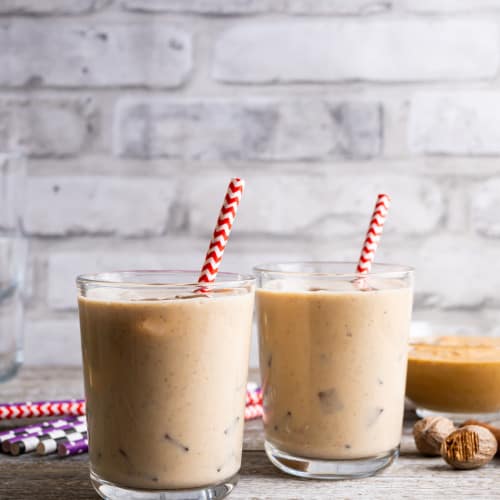 Two glasses of homemade peanut punch over ice sits on a wooden table. In each glass is a red and white paper straw. Whole nutmeg is beside the glasses and a bowl of peanut butter is in the background.