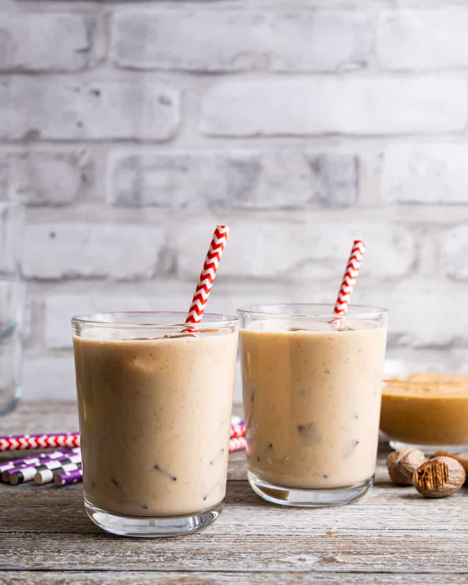 Two glasses of homemade peanut punch over ice sits on a wooden table. In each glass is a red and white paper straw. Whole nutmeg is beside the glasses and a bowl of peanut butter is in the background.