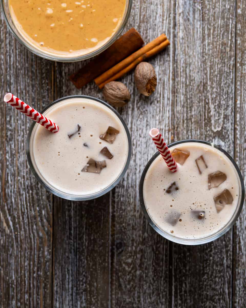 An overhead shot of two glasses of peanut punch with ice. In each glass is a red & white straw. Above the glasses is a bowl of peanut butter, cinnamon sticks and whole nutmeg.
