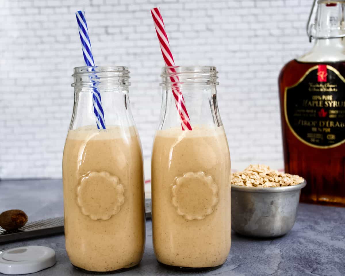 Two glass bottles filled with homemade peanut punch. A straw is in each bottle. A bottle of maple syrup, a small bowl of toasted oats and a whole nutmeg are in the background.