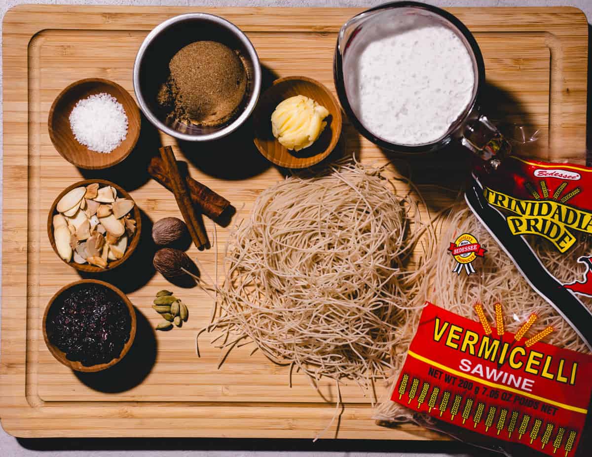 An overhead shot of ingredients for sawine on a wooden board: vermicelli, ghee, coconut milk, toasted almonds, currants, cinnamon, nutmeg, cardamom pod and brown sugar.
