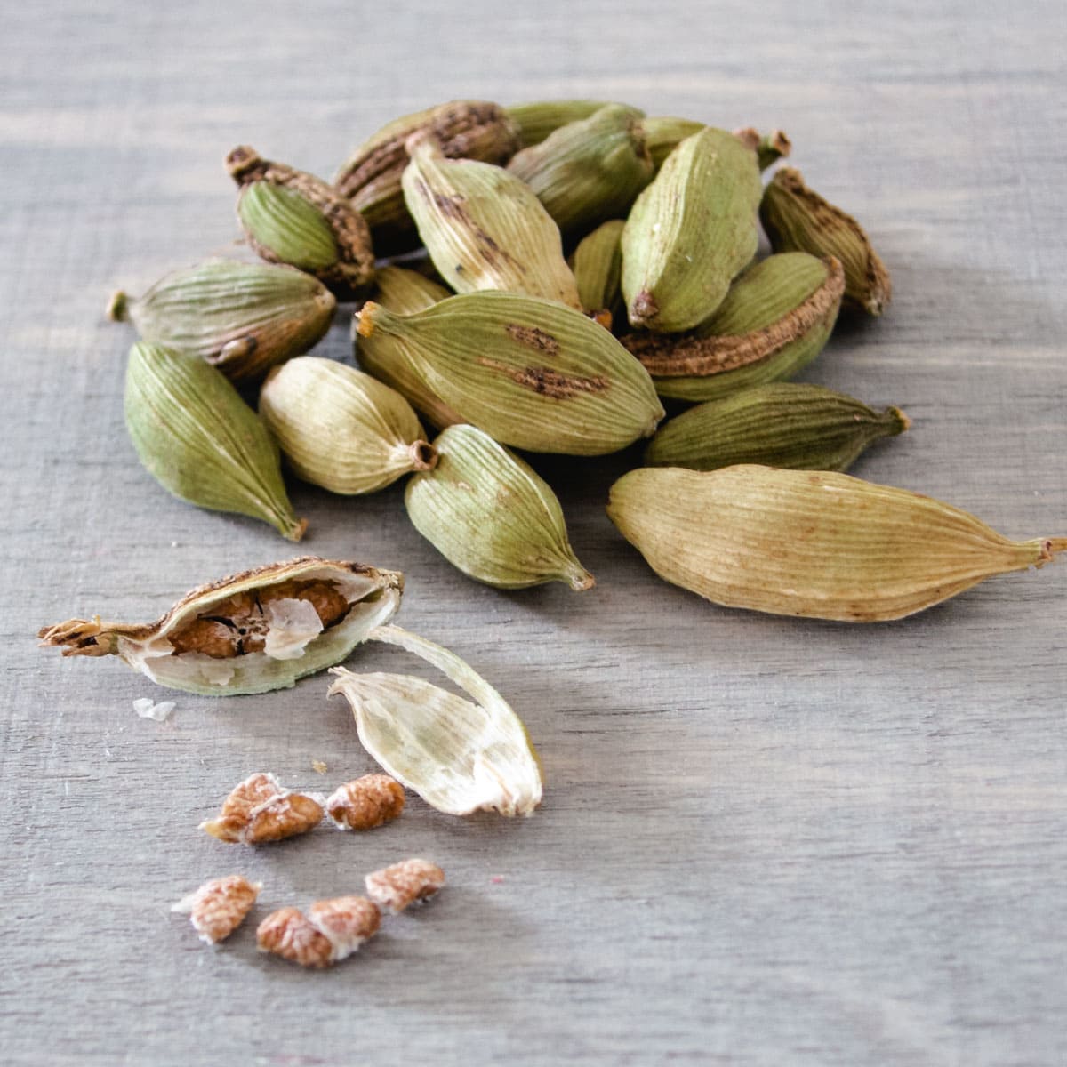 A small pile of green cardamom pods on a light grey surface. One cardamom pod is open and light brown seeds are spilling out.